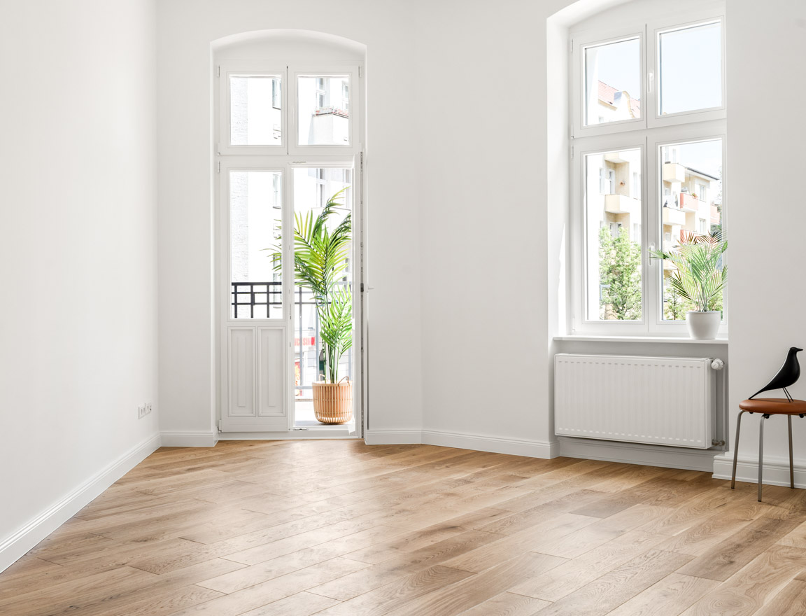Empty, bright room with wooden flooring, white walls, large windows and balcony access, plus two indoor plants.