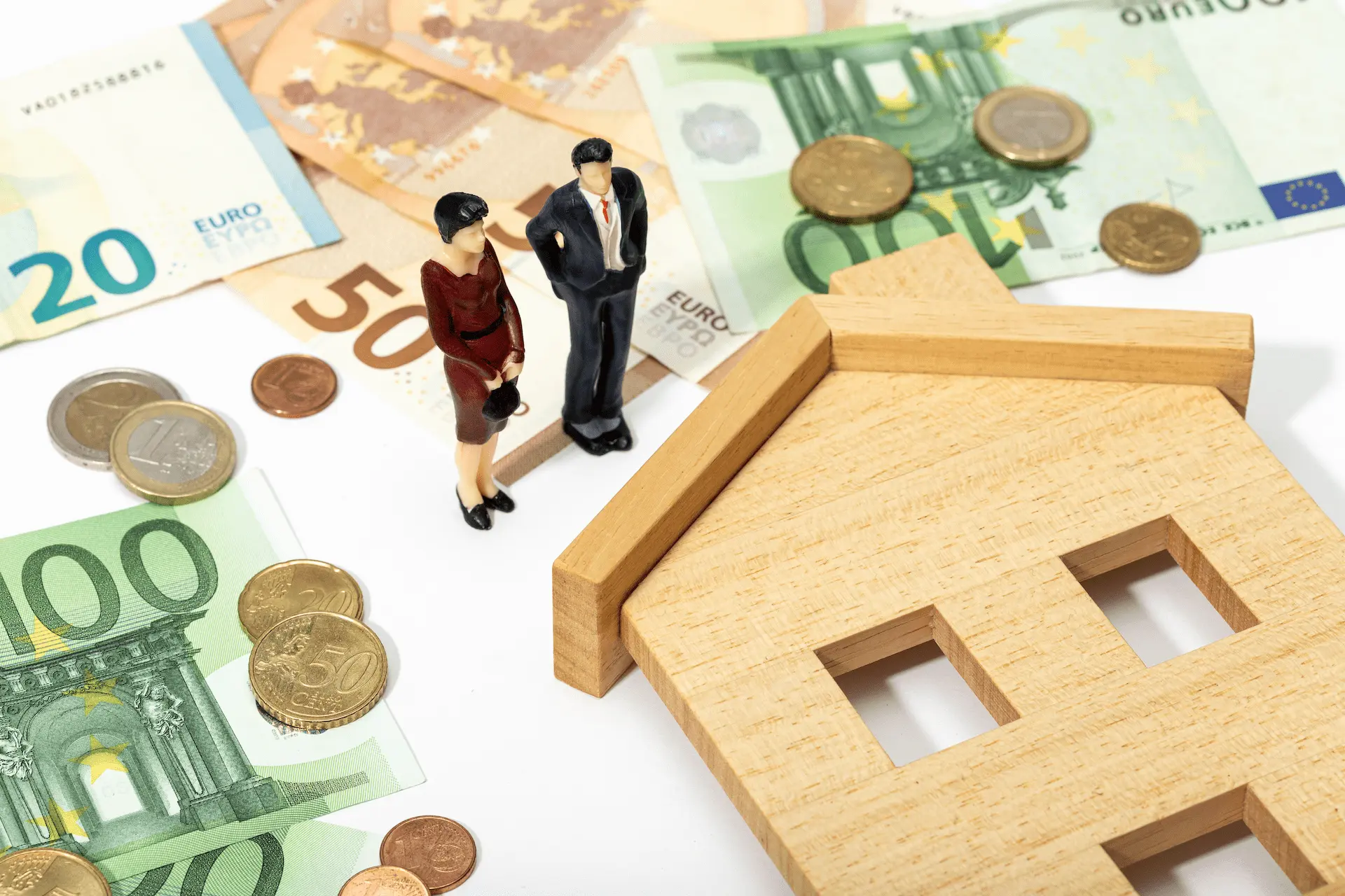 Miniature figures of a couple next to a wooden house model and euro banknotes symbolizing home purchase and subsidies.