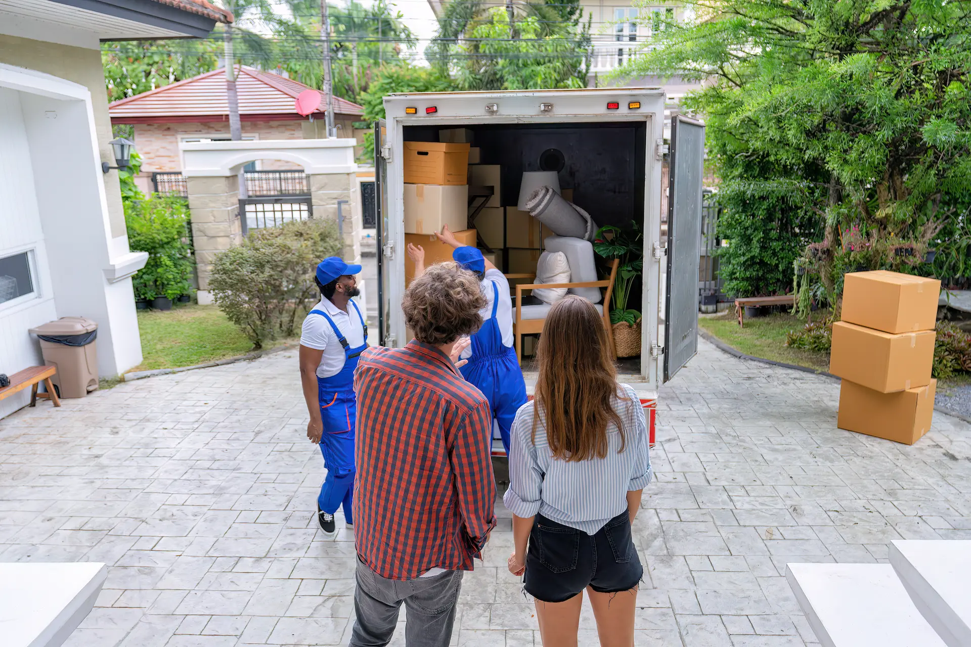 A couple watching two movers unloading furniture and boxes from a moving truck.