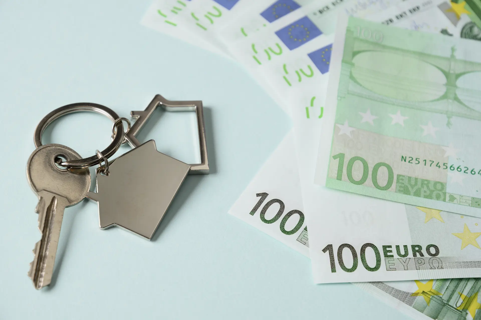 Keychain with a house-shaped keyring next to several 100-euro banknotes on a light background.