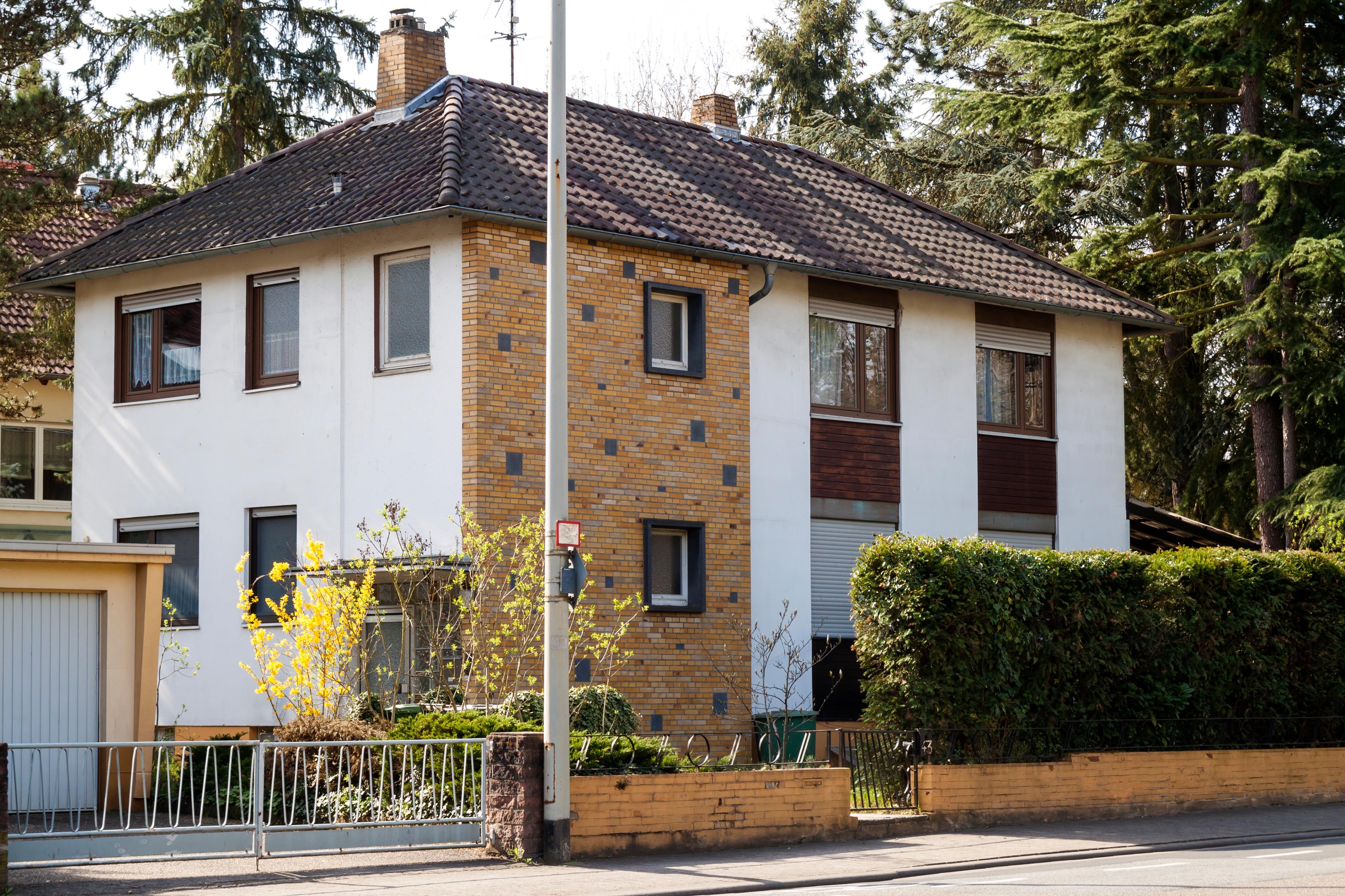 Two-story residential house with a mixed facade of white walls and brickwork, surrounded by trees and hedges.