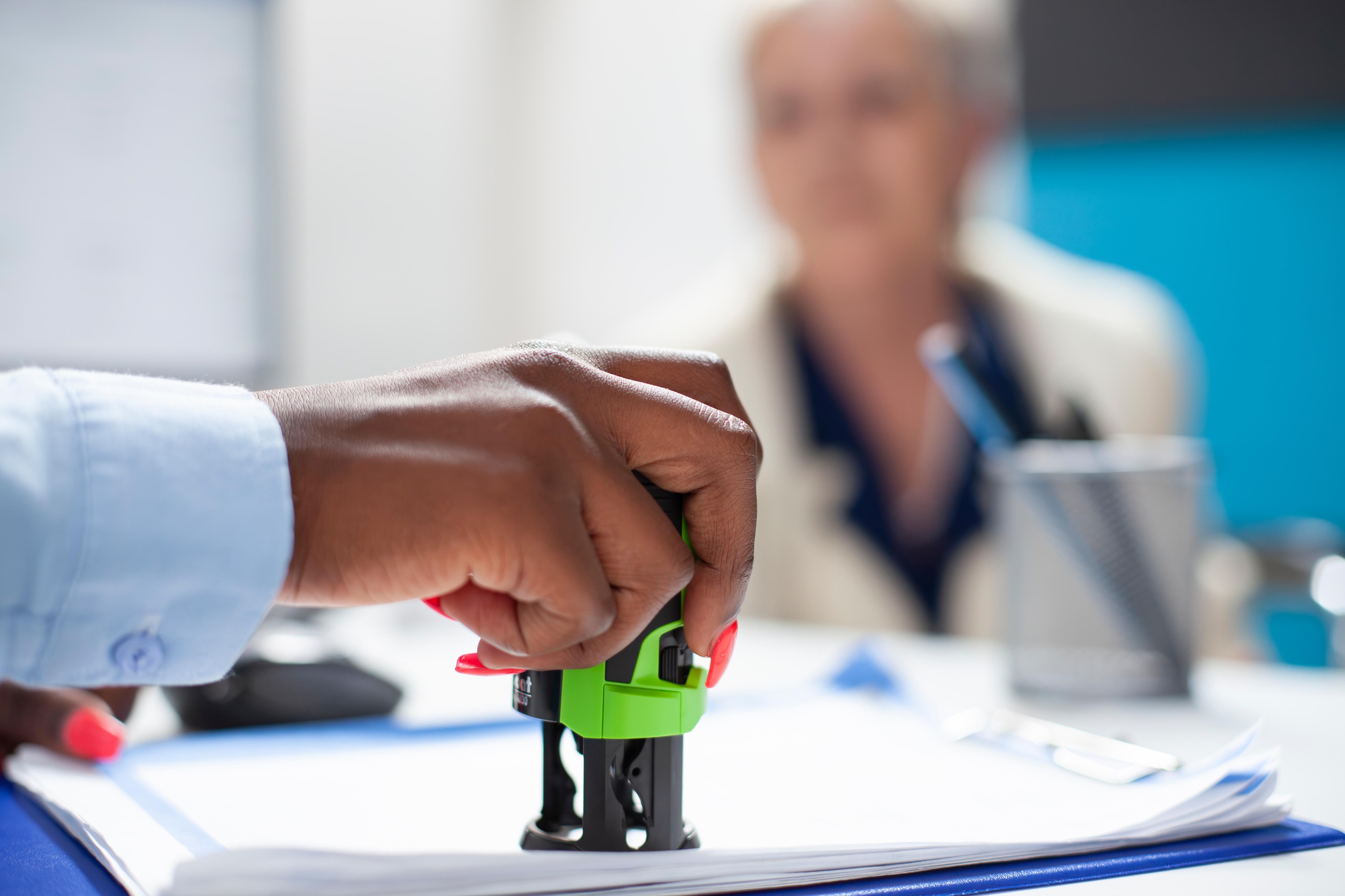Hand pressing a green stamp on a document, with a blurred person in the background.