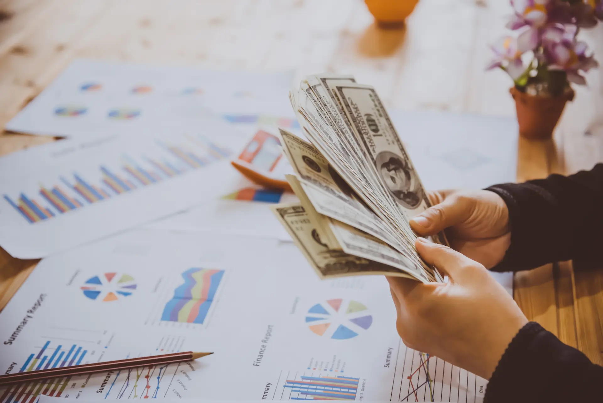 Person counting banknotes over spread financial charts on a table.
