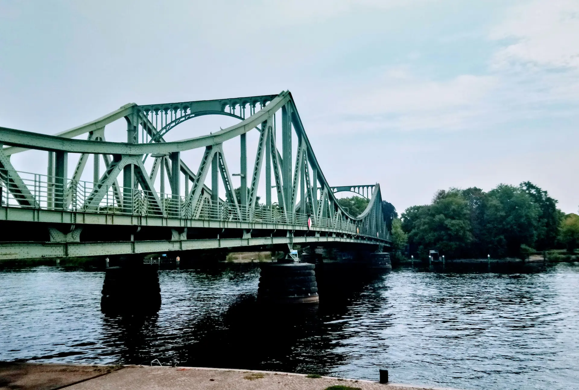 Green steel bridge spanning a calm river, surrounded by trees.