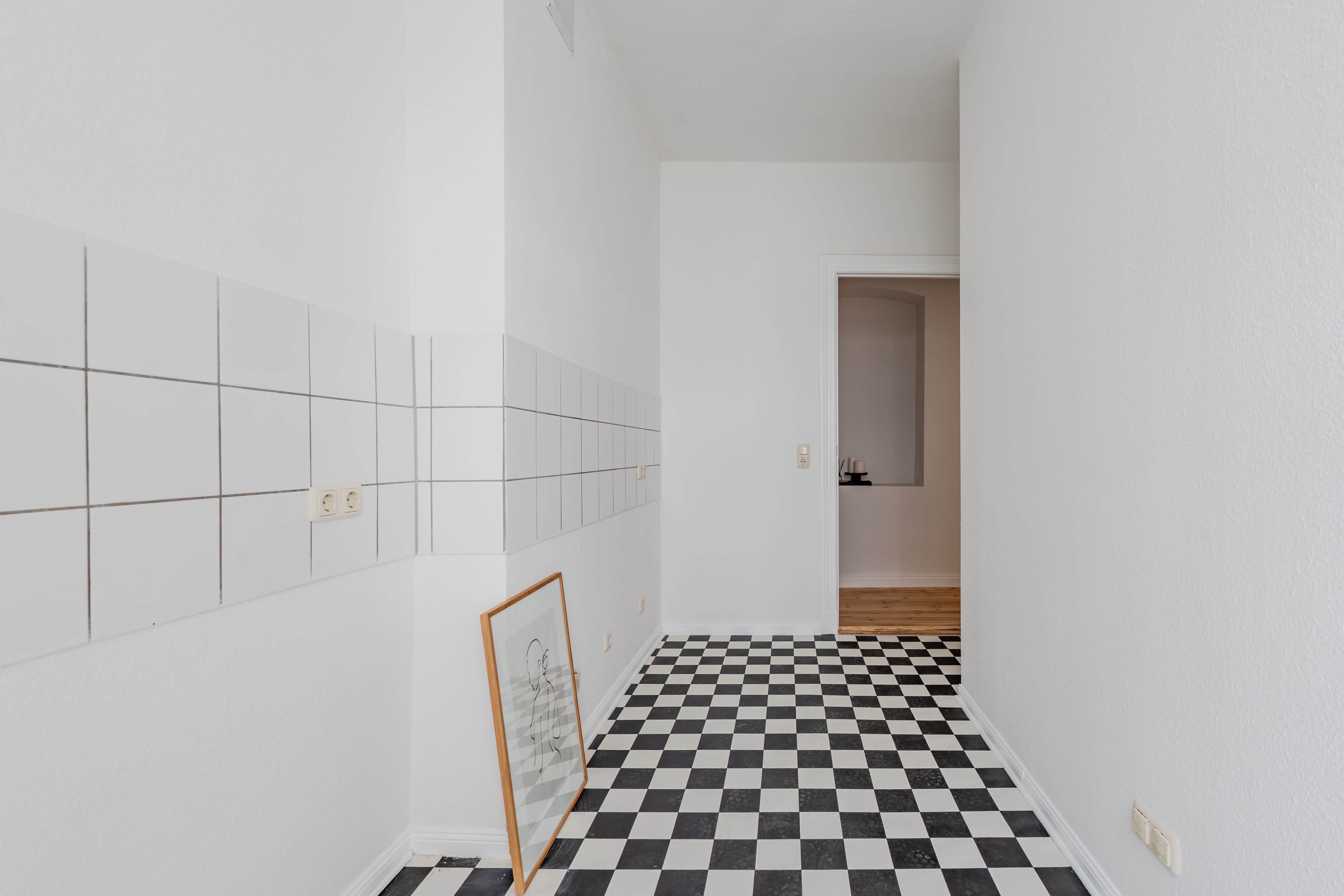 Leere Küche einer Altbauwohnung mit schwarz-weißem Schachbrettboden, weißen Wandfliesen und Blick in den angrenzenden Wohnbereich