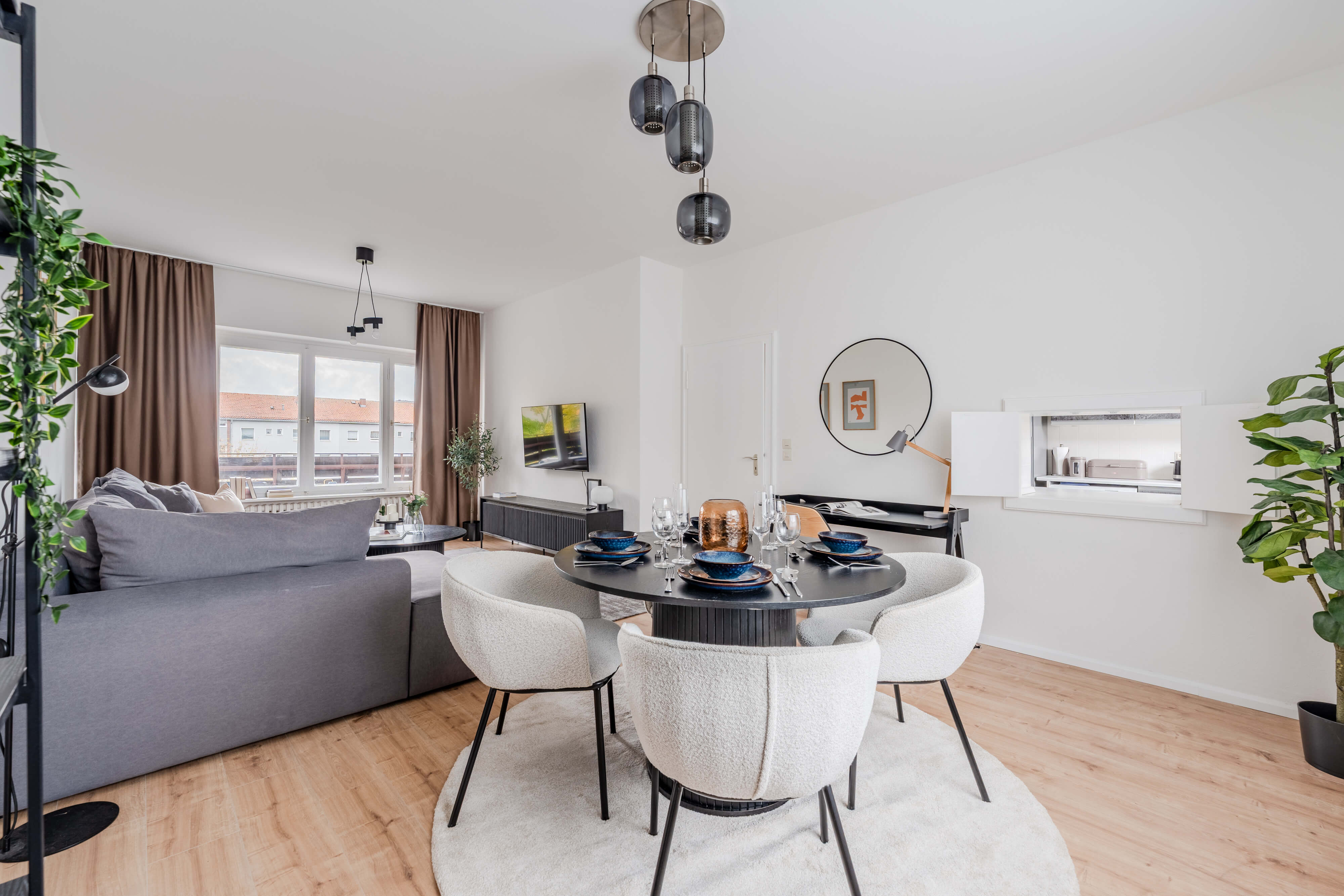 Offenes Wohn- und Esszimmer mit rundem Esstisch, weißen Polsterstühlen, grauem Sofa, großen Fenstern und stilvollen Dekoelementen.