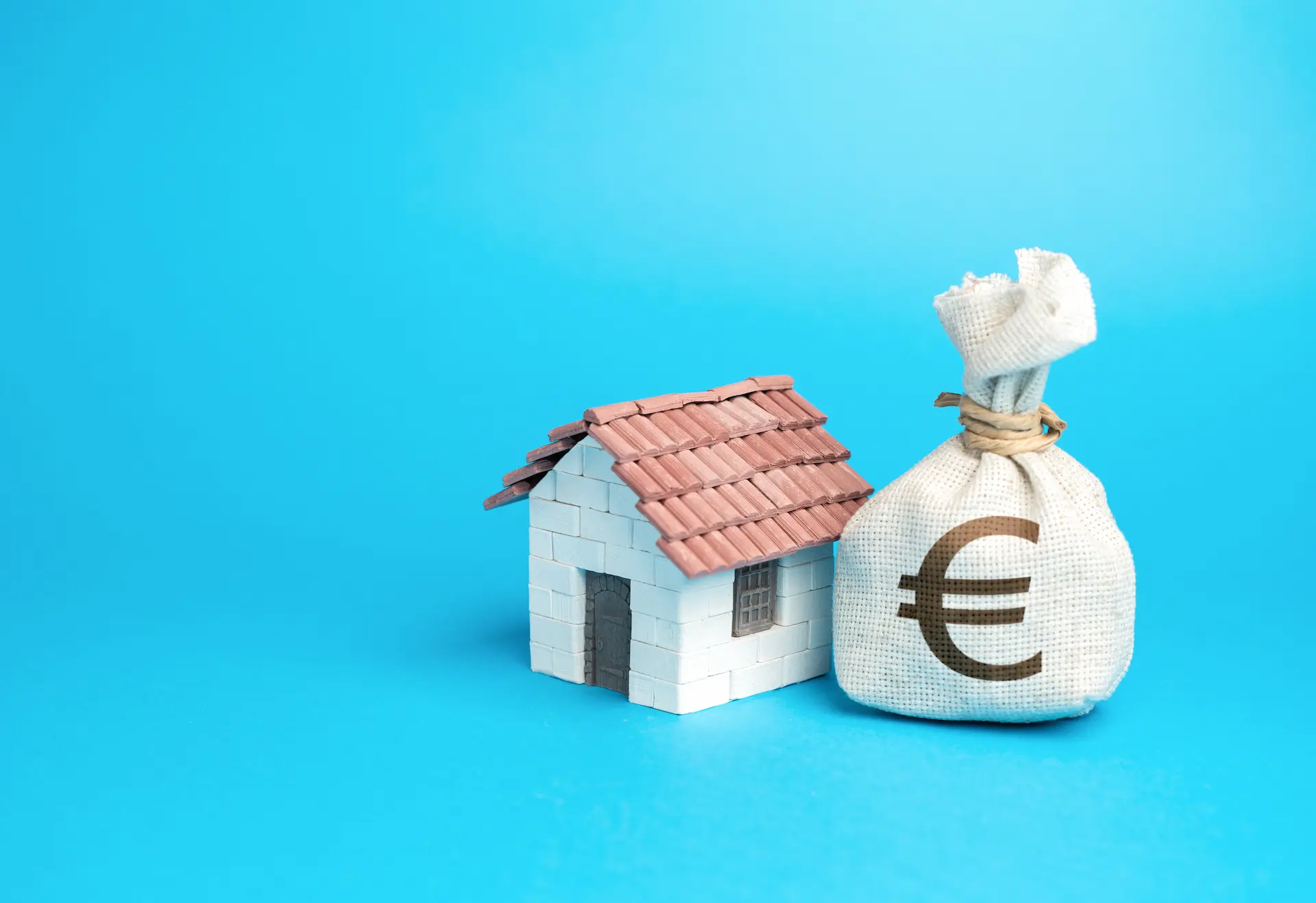 Miniature house next to a money bag with euro symbol on a blue background.