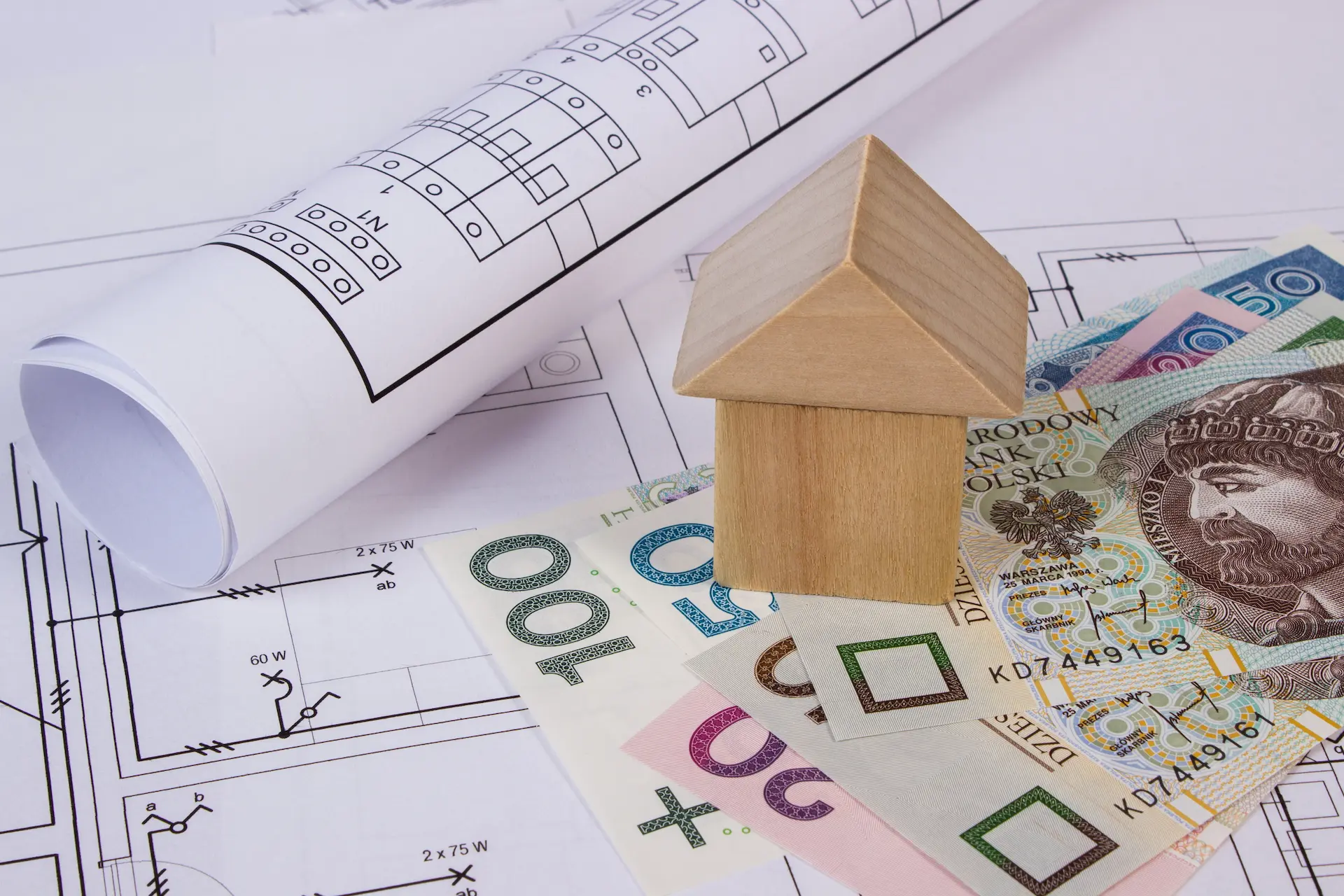 Wooden house model on blueprints next to banknotes, symbolizing real estate financing.