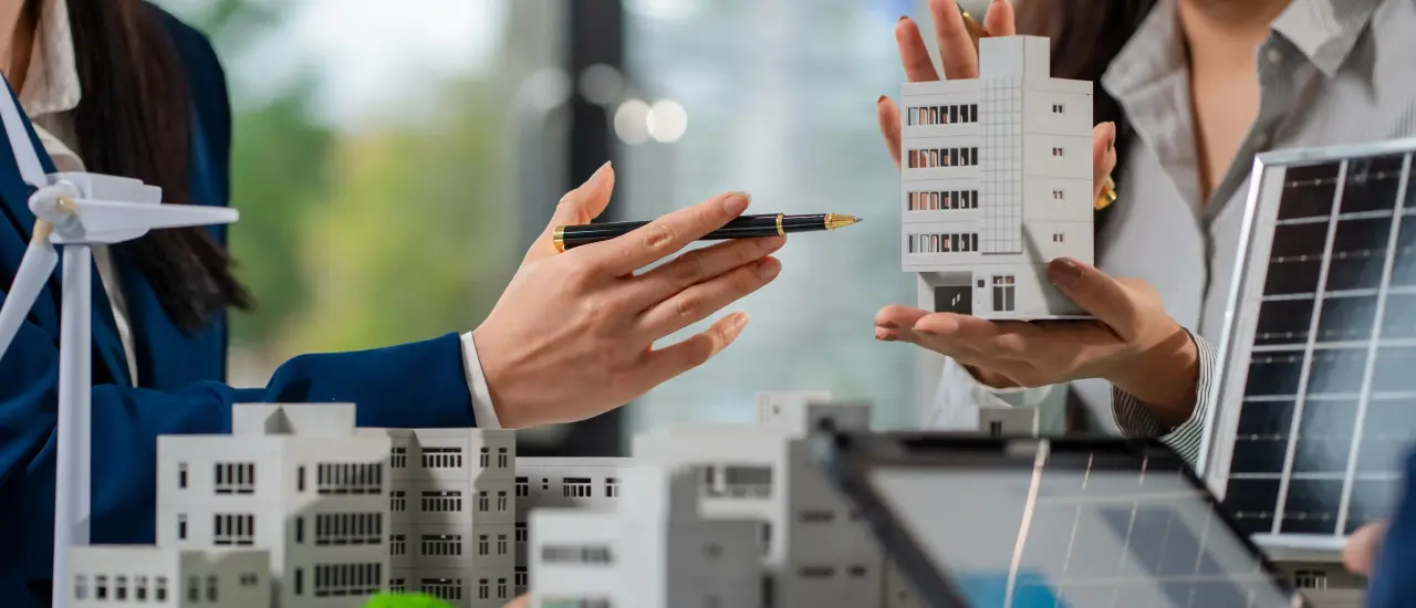 Geschäftsfrauen diskutieren über ein Modellgebäude, im Vordergrund Solarmodul und Windrad.