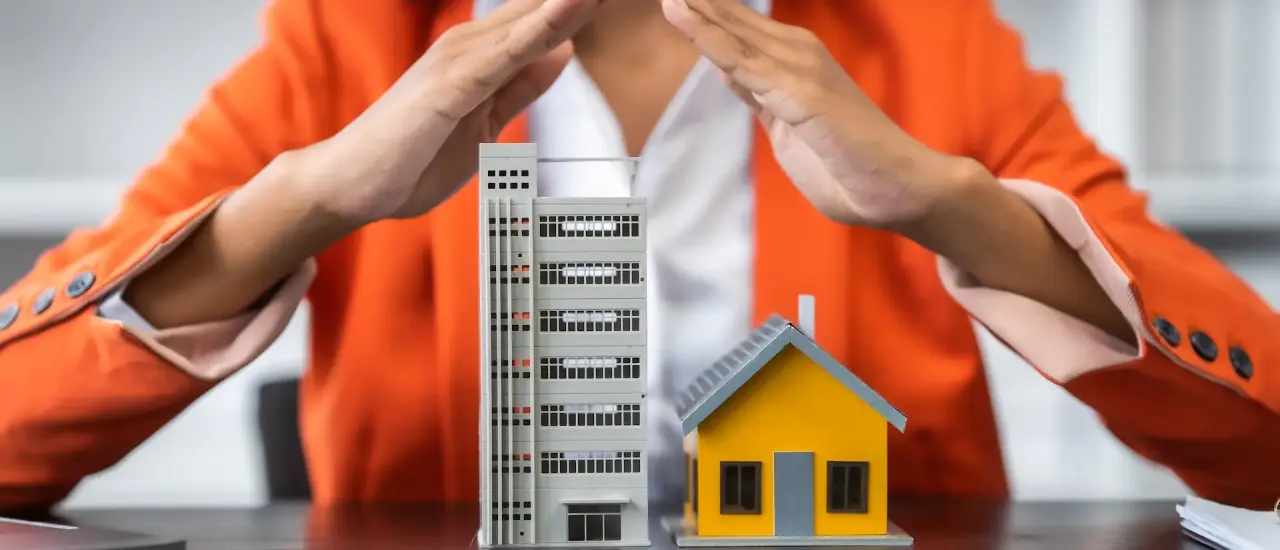 Frau schützt Modelle von Haus und Gebäude mit Händen – Symbolbild für Haftpflicht- und Hausratversicherung