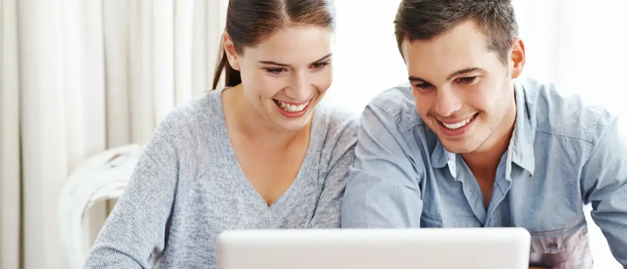 Junges Paar sitzt lächelnd vor einem Laptop und betrachtet gemeinsam Inhalte auf dem Bildschirm.
