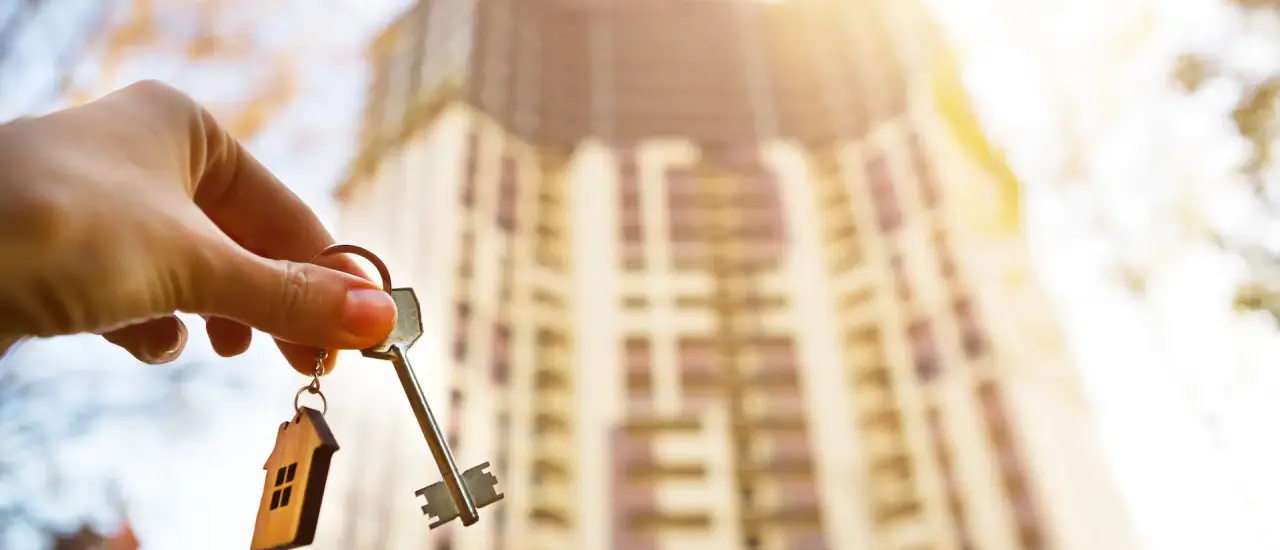Hand hält Wohnungsschlüssel vor einem unscharfen Mehrfamilienhaus