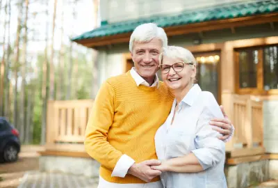 Lächelndes älteres Paar steht umarmt vor einem modernen Haus im Wald.