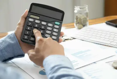 Hände halten Taschenrechner über Finanzunterlagen und Münzen.