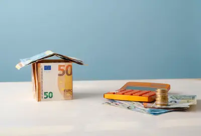 Haus aus 50-Euro-Schein neben Taschenrechner und Münzstapeln.