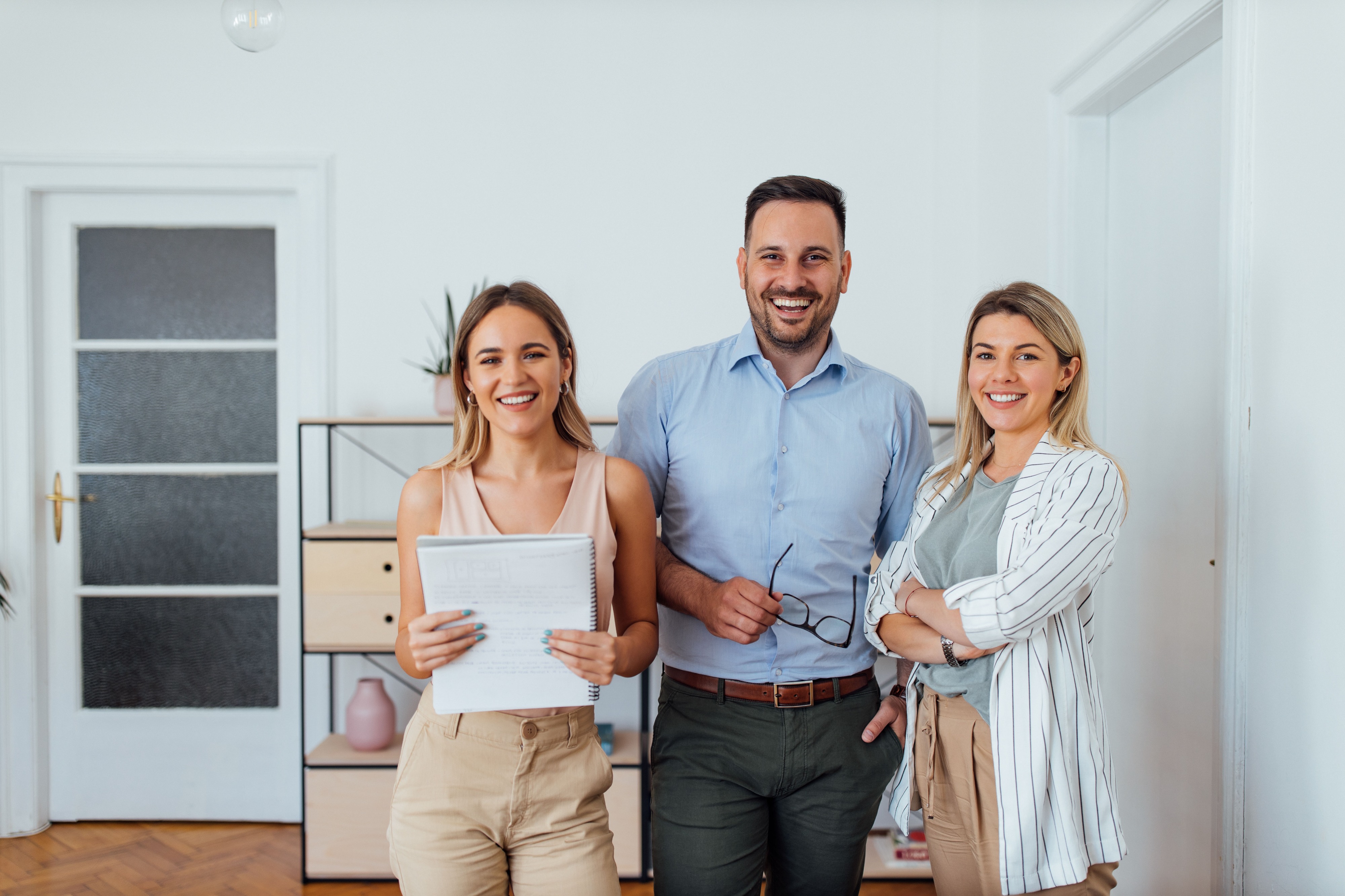 Drei lächelnde Immobilienmakler stehen in einem modernen Büro. Eine Frau hält Unterlagen in den Händen, ein Mann in Hemd und Hose hält seine Brille, eine weitere Frau trägt ein gestreiftes Jacket. Im Hintergrund ein Regal mit Deko.