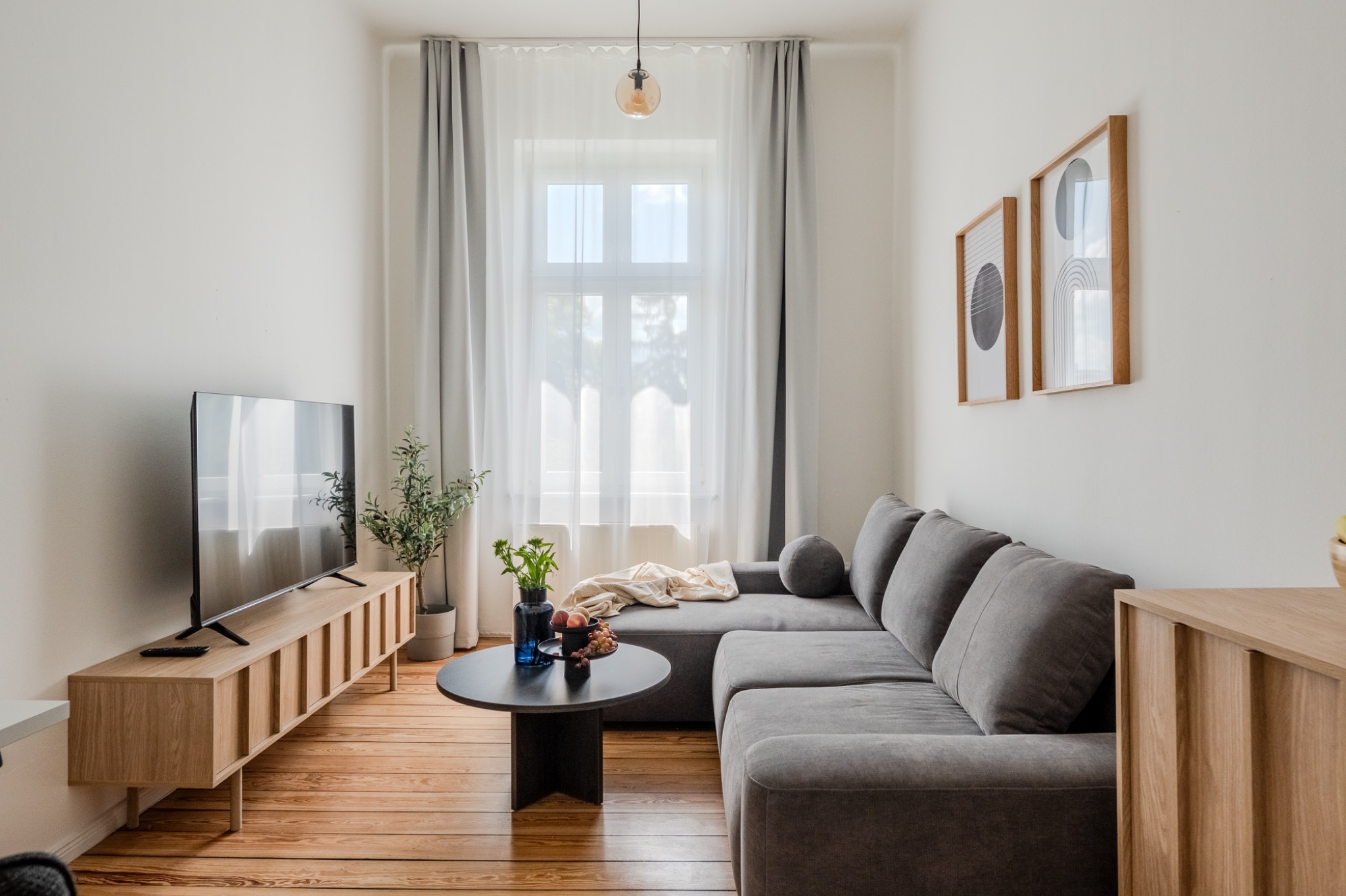 Bright, modern living room with a grey corner sofa, light curtains, wooden floor, TV on a sideboard, and minimalist wall decor.