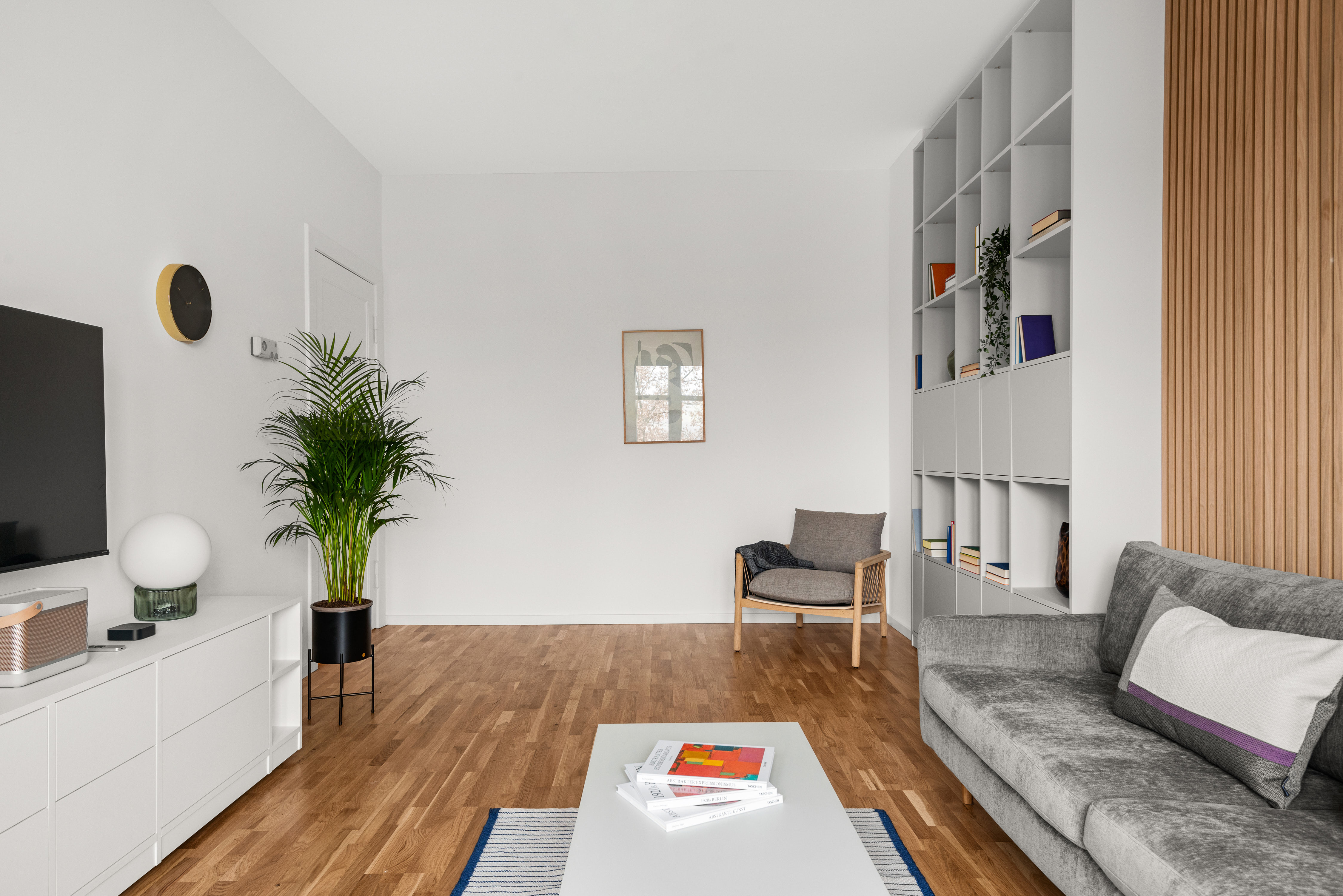 Modern living room with a grey sofa, large shelving unit, indoor plant, wall art, and white TV sideboard on light wooden flooring.