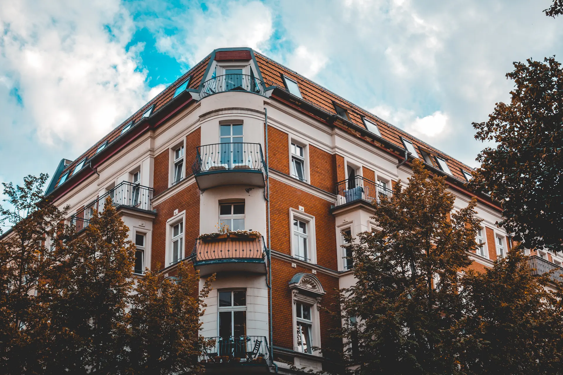 Historic corner apartment building with red brick façade, balconies, and trees.