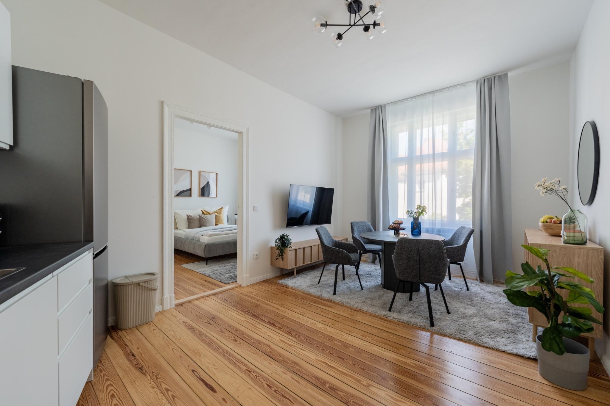 Open living and dining area with grey upholstered chairs, wall-mounted TV, large curtained windows, and view into a cozy bedroom with a double bed.