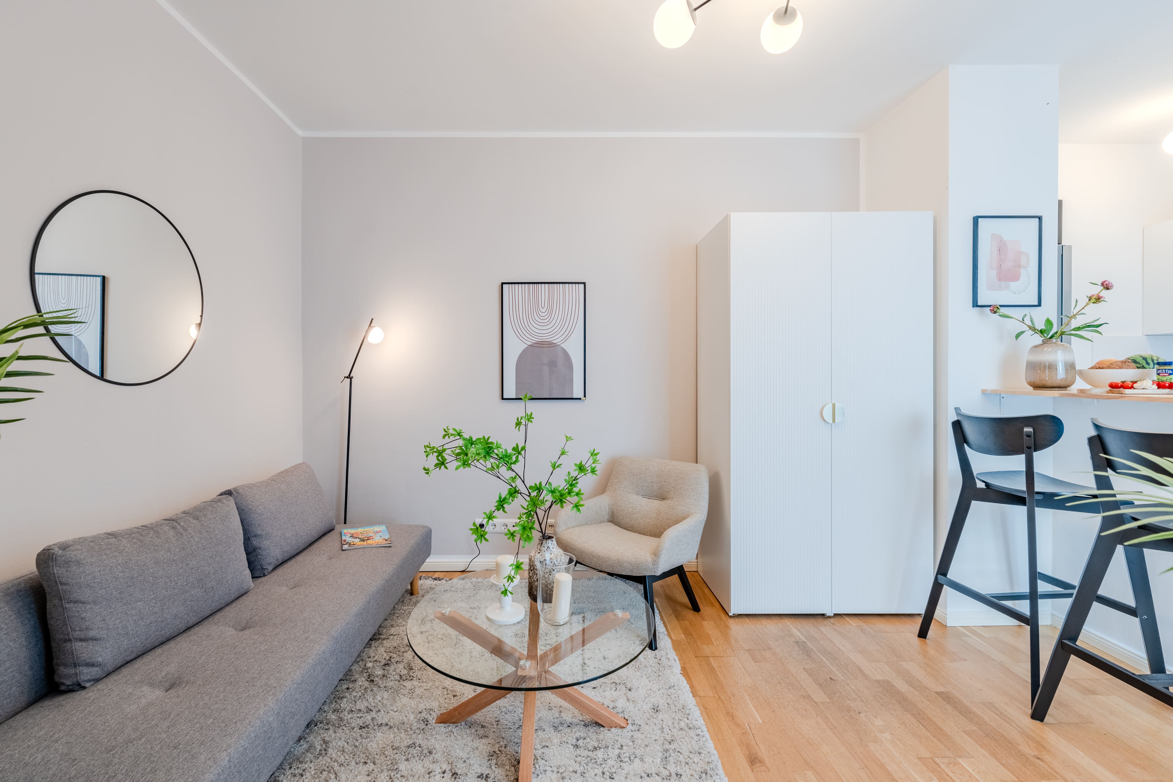Modern eingerichtetes Wohnzimmer mit grauem Sofa, gemütlichem Sessel, runder Glas-Couchtisch und offener Küche im Hintergrund.