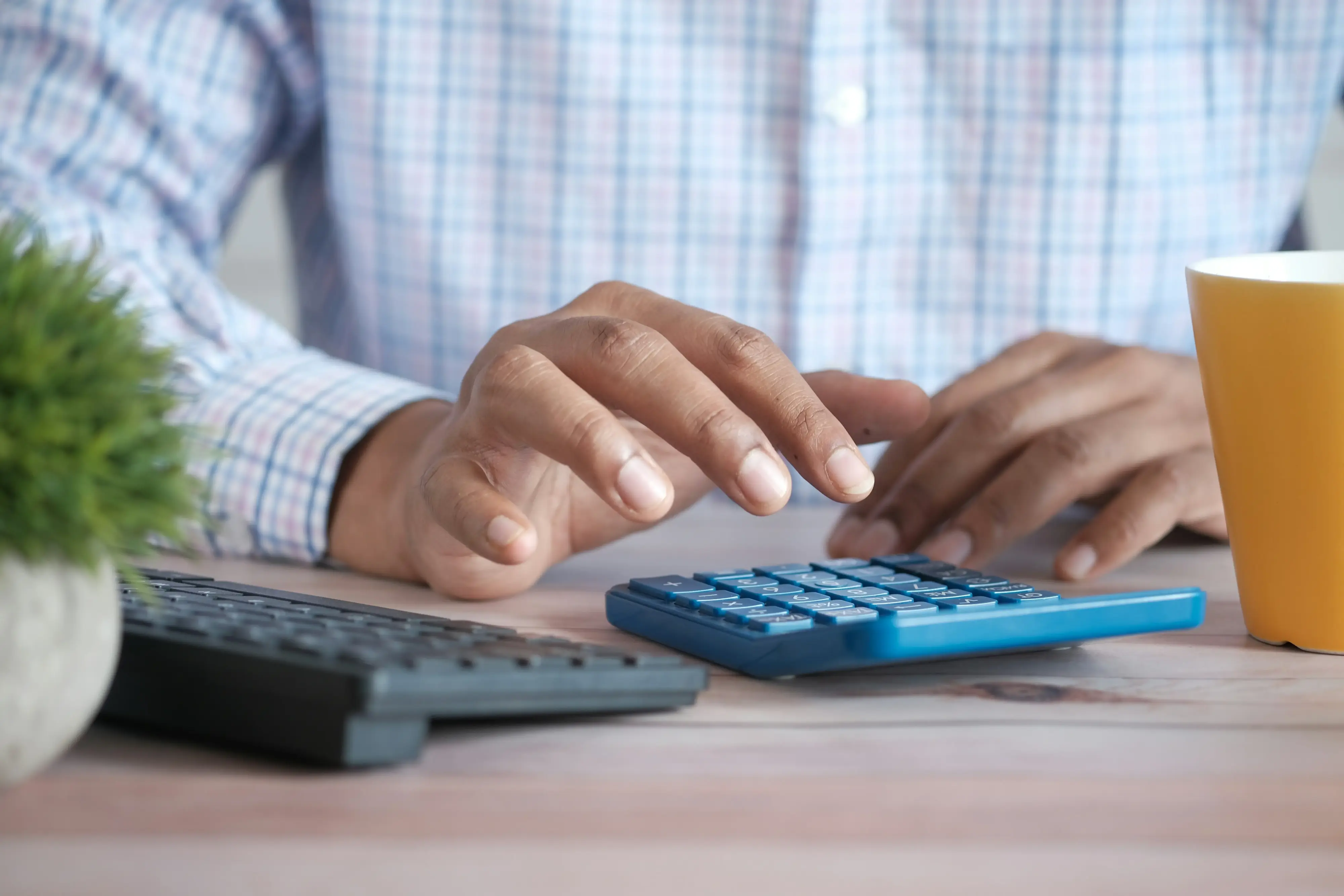 Person typing on a calculator at a desk next to a keyboard and mug.
