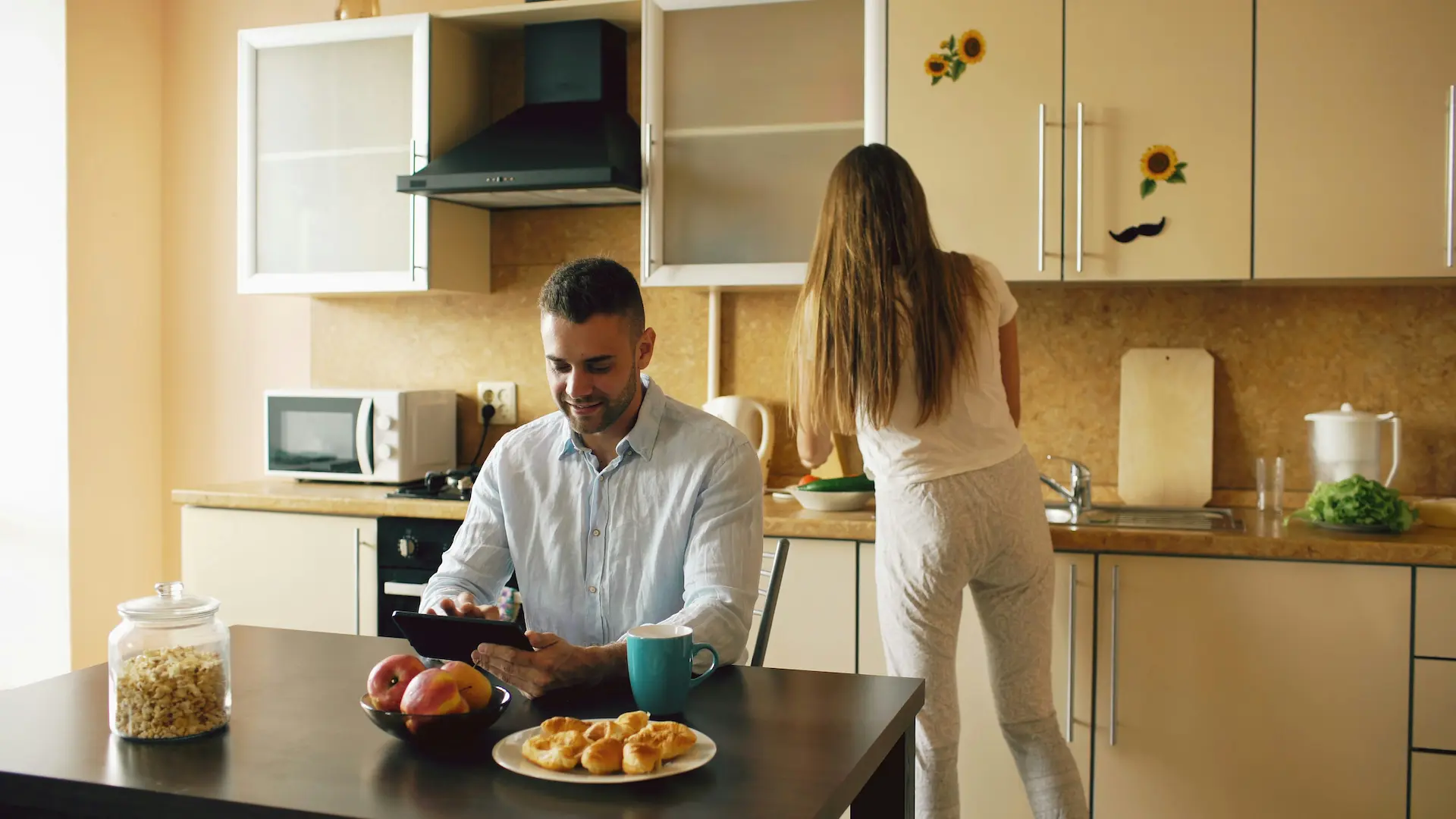 One person sits at a table with a tablet, another works in the kitchen