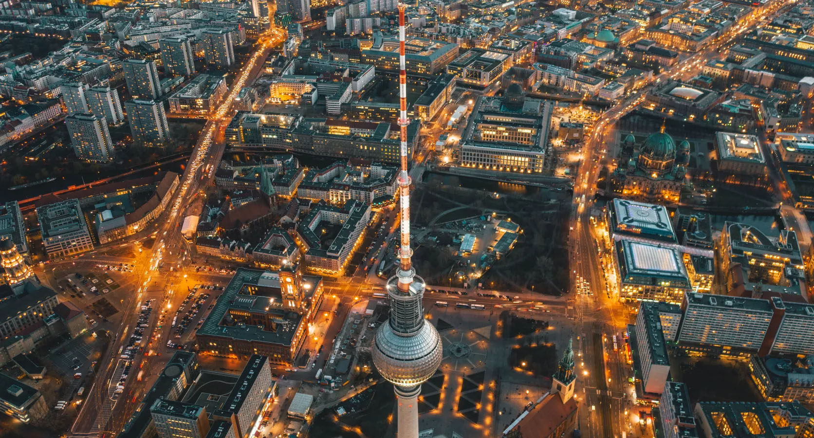 Luftaufnahme von Berlin bei Abenddämmerung mit beleuchtetem Fernsehturm und umliegenden Stadtteilen.