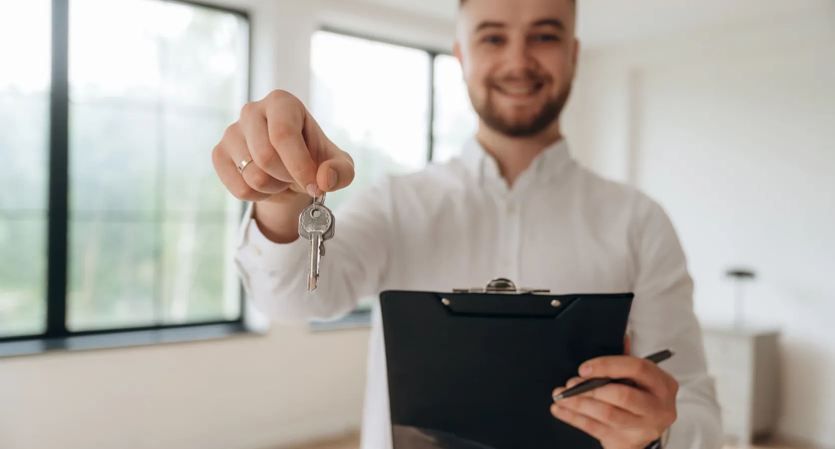 Lächelnder Immobilienmakler in heller Wohnung übergibt symbolisch einen Wohnungsschlüssel und hält dabei ein Klemmbrett in der Hand.