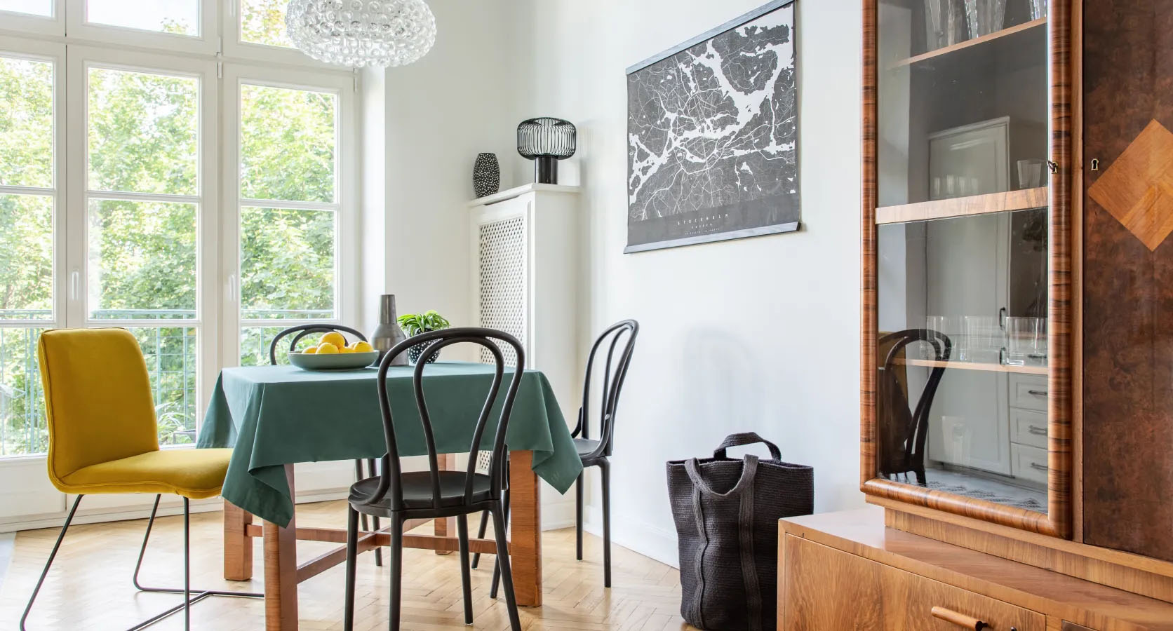 Helles Esszimmer mit großen Fenstern, einem Holztisch mit grüner Tischdecke, gemischten Stühlen, darunter ein gelber Polsterstuhl, und einem Vintage-Holzschrank mit Glasvitrine.